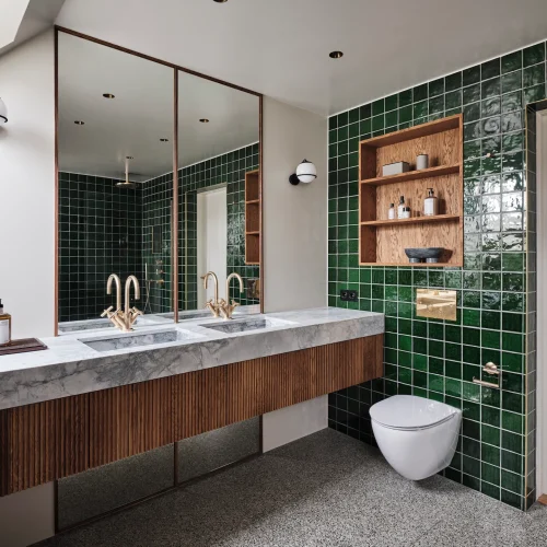 Moderne badeværelse med dobbelt håndvask i marmor, messingarmaturer, dybgrønne fliser og væghængt toilet.